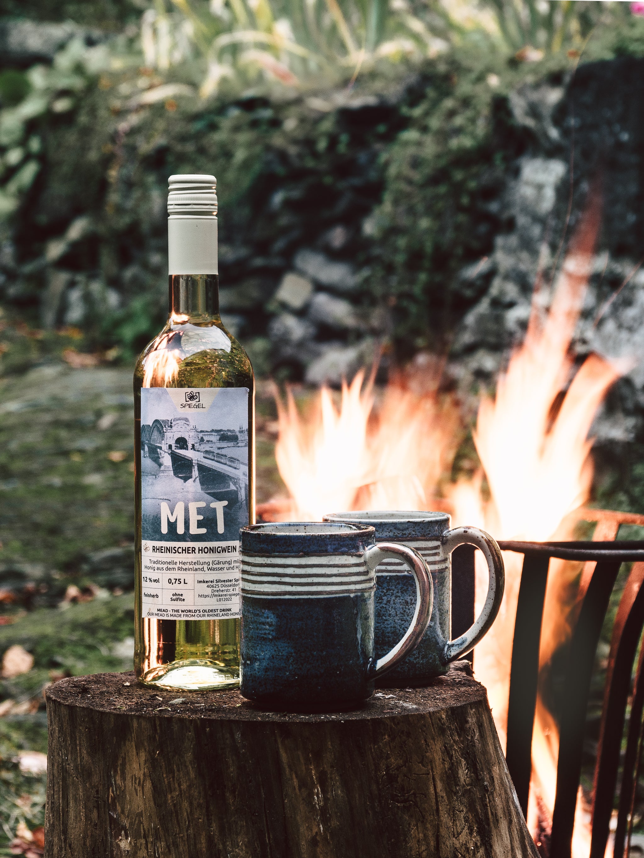 Flasche Rheinischer Honigwein mit zwei Steingutbechern vor Lagerfeuer, aufgenommen in der Natur – Met aus Düsseldorf.