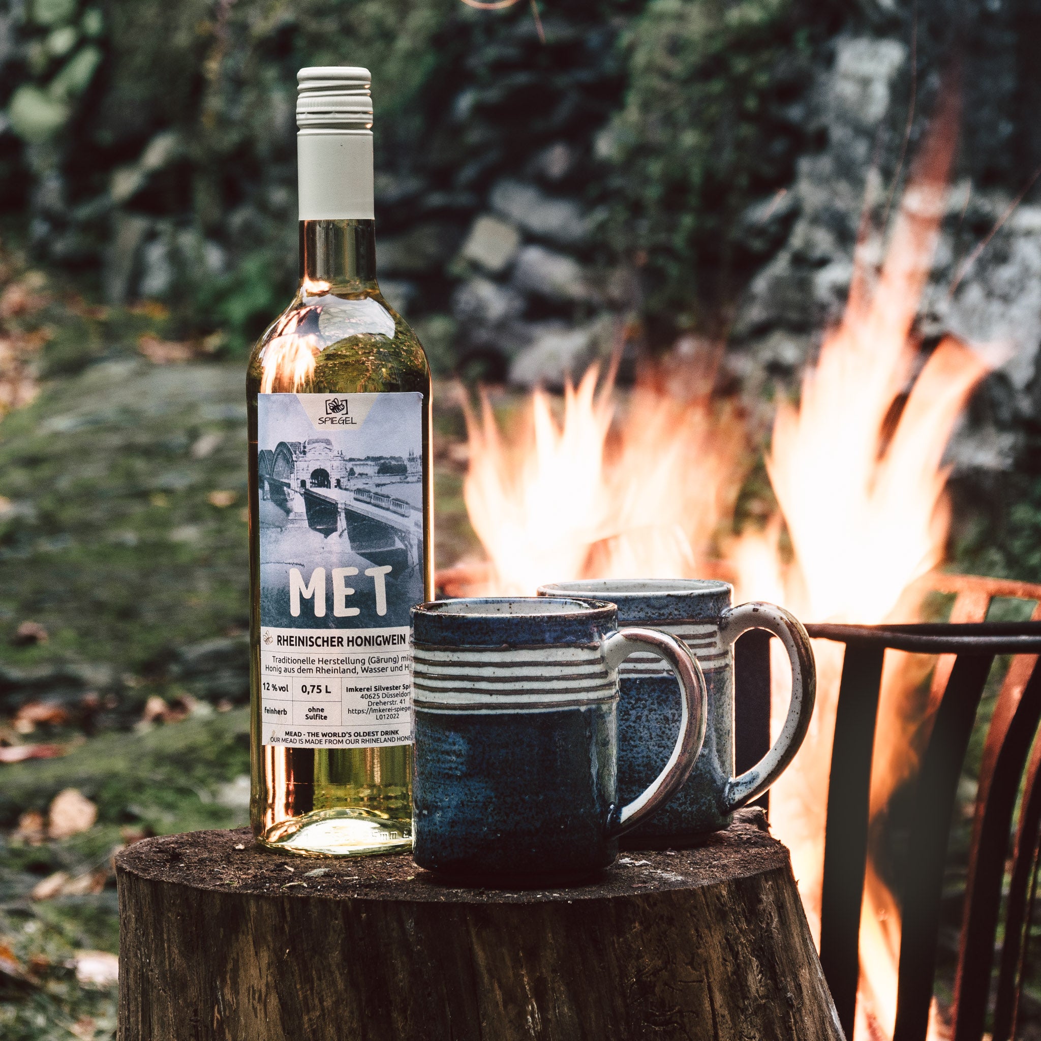 Flasche Rheinischer Honigwein mit zwei Steingutbechern vor Lagerfeuer, aufgenommen in der Natur – Met aus Düsseldorf.