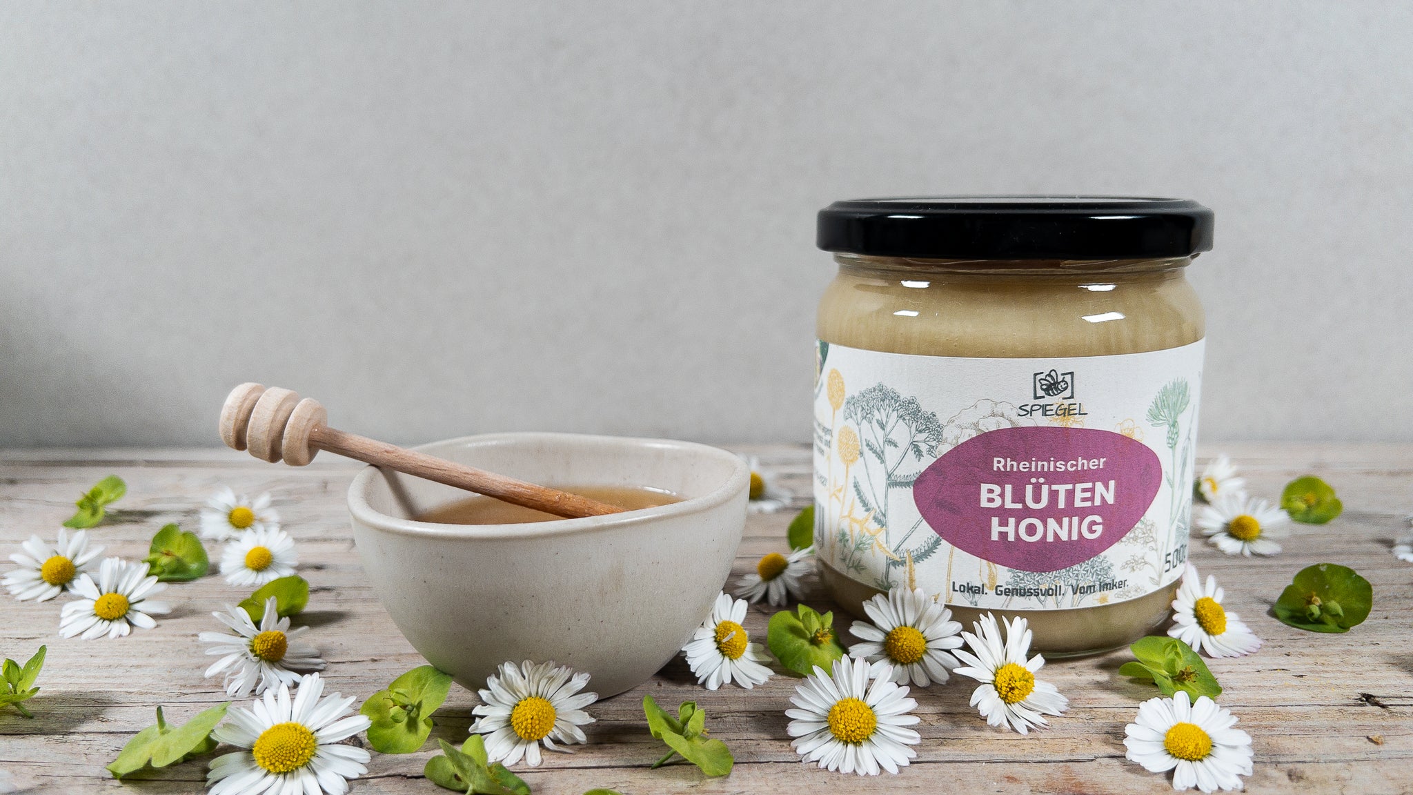 Glas Blütenhonig und Steingutschale mit Holzlöffel auf Holzplatte mit frischen Blüten.
