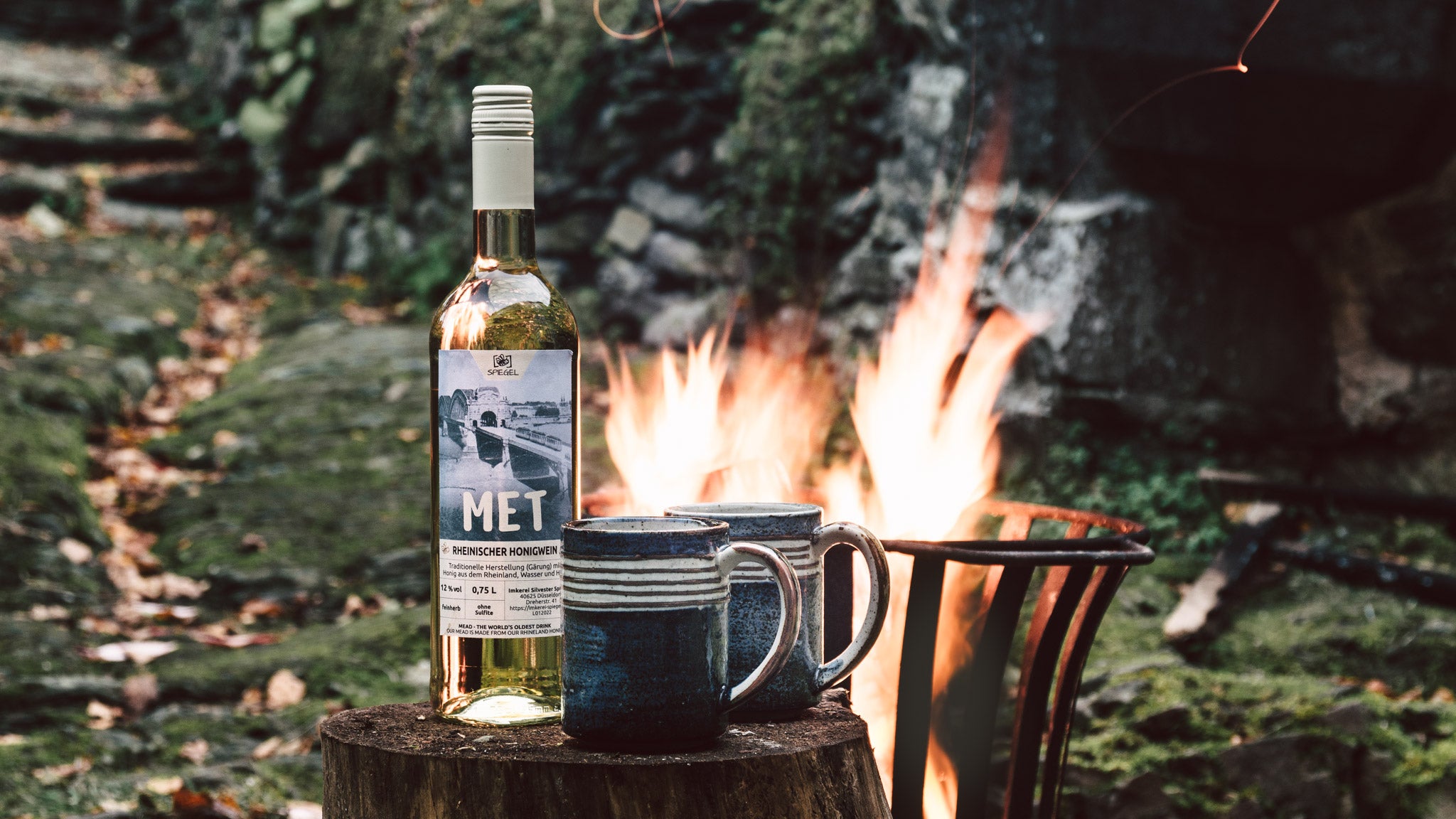 Flasche Rheinischer Honigwein mit zwei Steingutbechern vor Lagerfeuer, aufgenommen in der Natur – Met aus Düsseldorf.